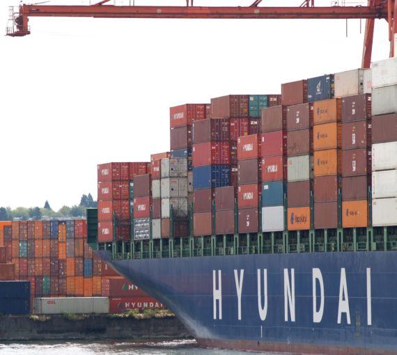 Shipping containers stacked near a port. Blue Hyundai tanker is in the foreview.